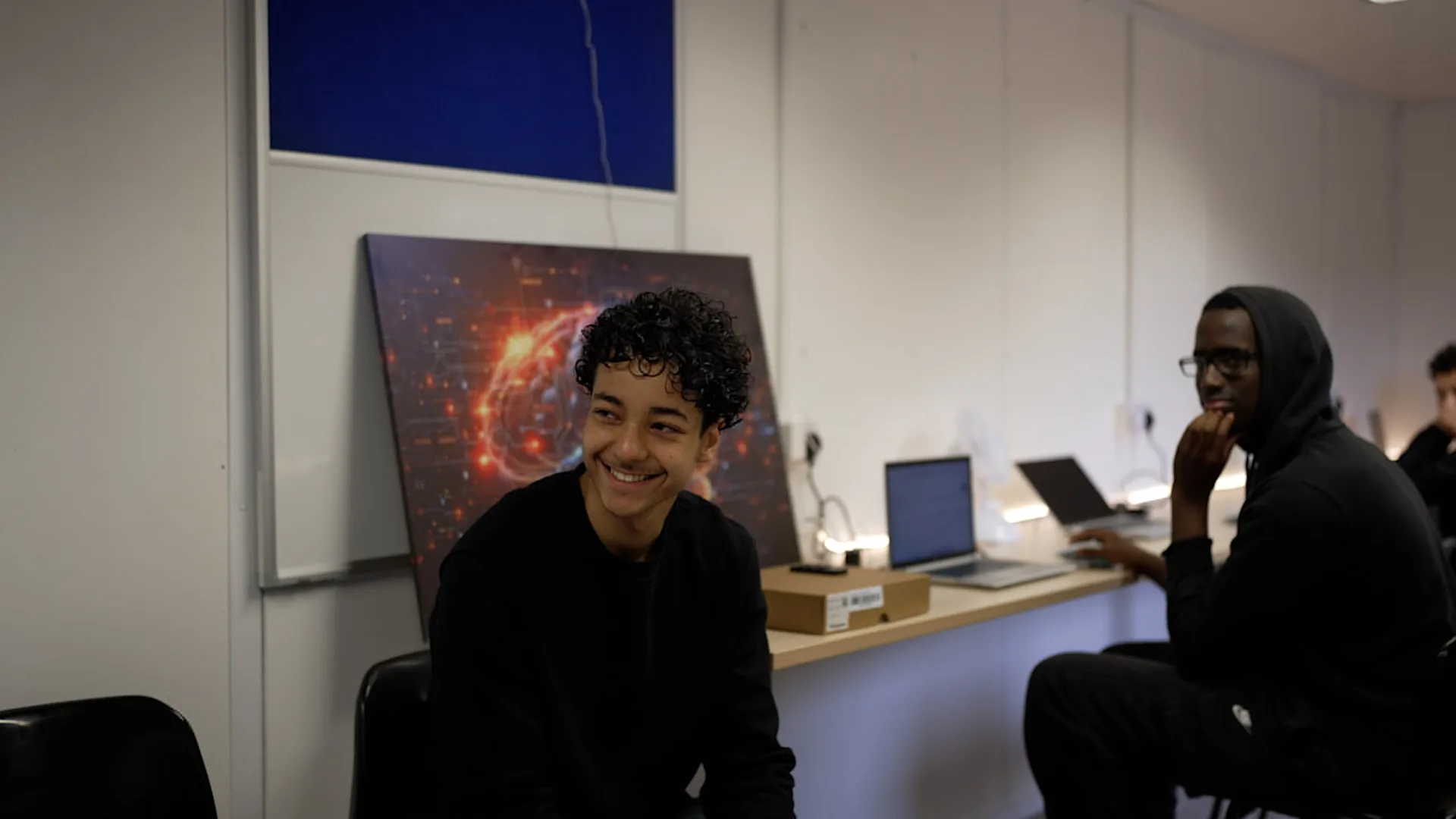 A teenager smiling in the Hikora classroom, a second young person at a laptop behind him