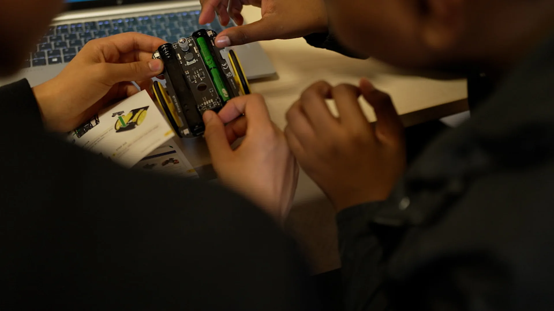 Two pairs of hands fitting a battery into a small wheeled robot, its build instructions open on the desk