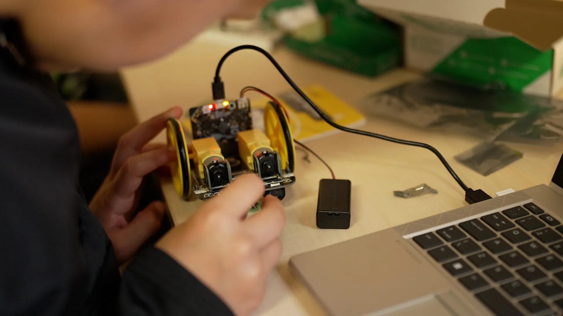 Hands fitting a motor to a small wheeled robot, its board lit up and cabled to a laptop