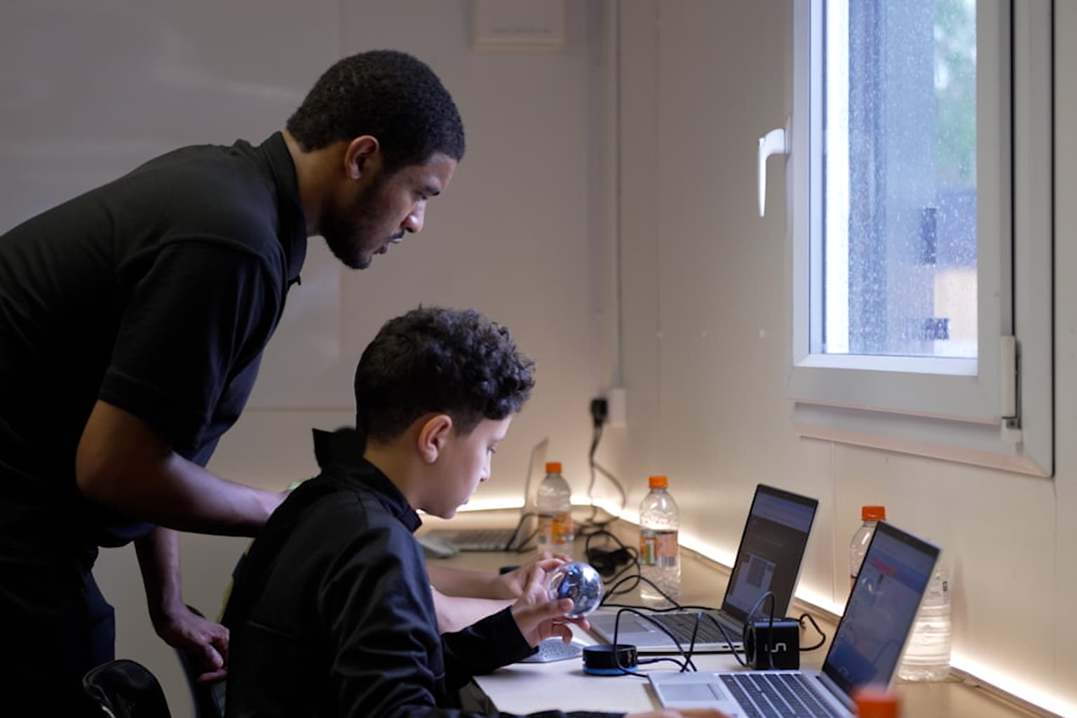 An adult in a black polo shirt leaning over a boy who is holding a Sphero robot at a laptop, by a window