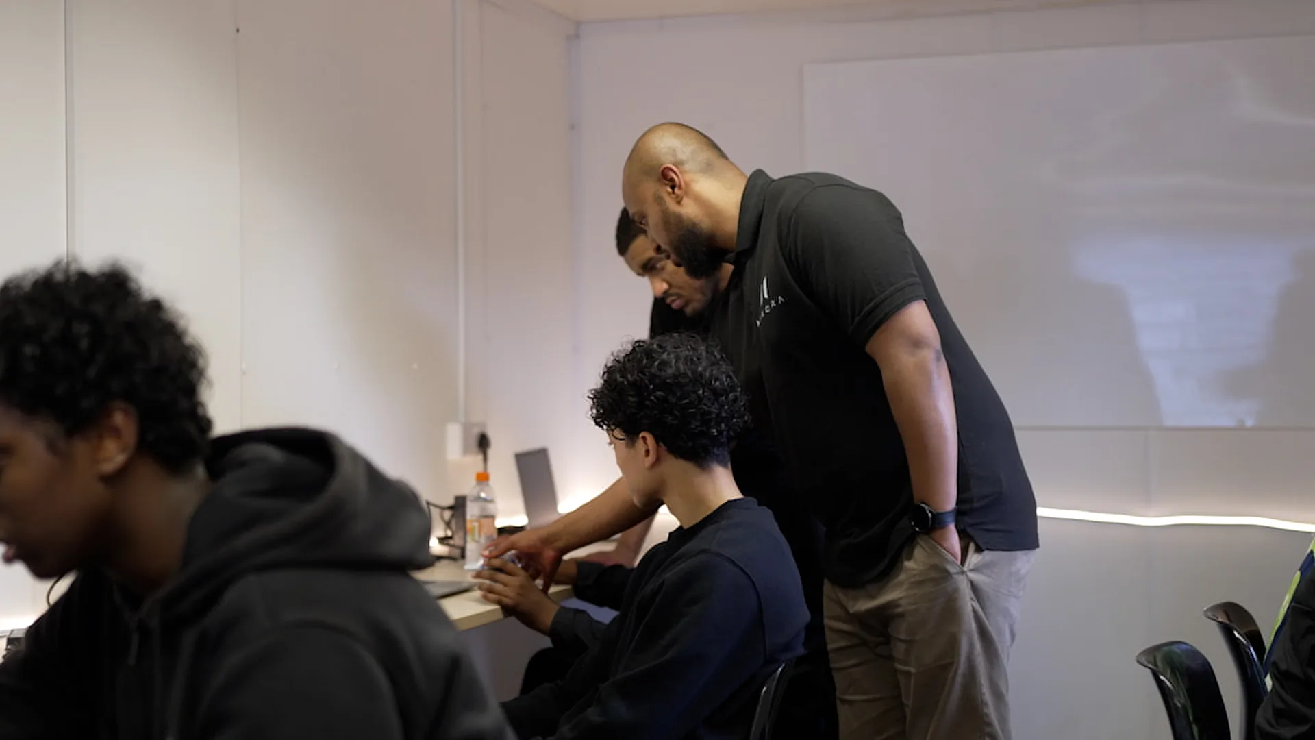 Two adults leaning in over a teenager working at a laptop, with a second teenager in the foreground