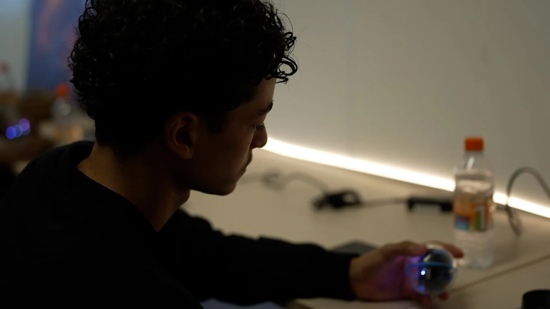 A teenager in profile holding a glowing Sphero robot at a desk, lit by an LED strip along the wall