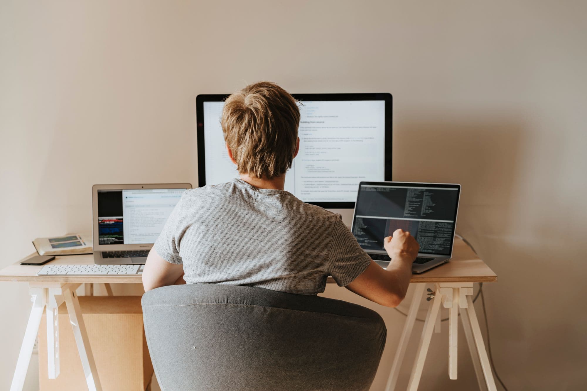 A young person coding across a desktop monitor and laptops