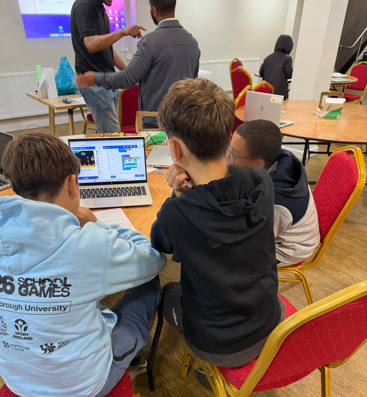 Three boys leaning over a laptop running block-based code at a workshop table, a small robot and printed worksheets beside them, an adult pointing at a projector screen behind and more tables across the room
